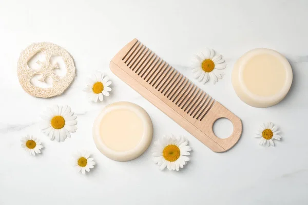 Solid shampoo bars, comb and chamomiles on white marble table, flat lay. Hair care