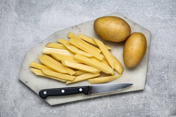 Whole and cut raw potatoes with knife on light grey table, top view. Cooking delicious French fries