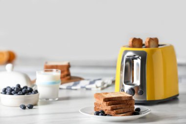 Kızarmış ekmek dilimleri, yaban mersini ve beyaz mermer masada bir bardak sütle birlikte sarı tost makinesi.