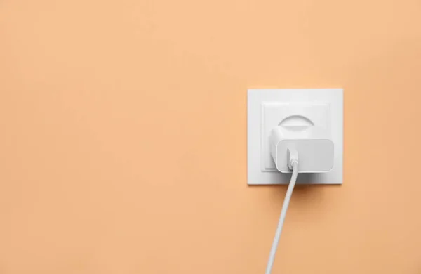 Charger adapter plugged into power socket on pale orange wall, space for text. Electrical supply