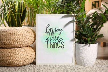 Frame with words Enjoy The Little Things near houseplants on carpet