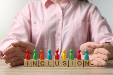 Woman protecting colorful pawns and wooden cubes with word Inclusion at table, closeup
