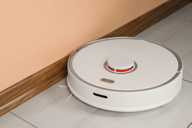 Beyaz fayanslı zeminde, bej duvarın yanında robotik süpürge var.