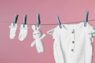 Farklı bebek kıyafetleri ve pembe arka planda çamaşır ipinde kuruyan tavşan oyuncağı.