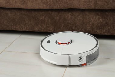 Robotik süpürge beyaz fayanslı zeminde kapalı koltuğun yanında.