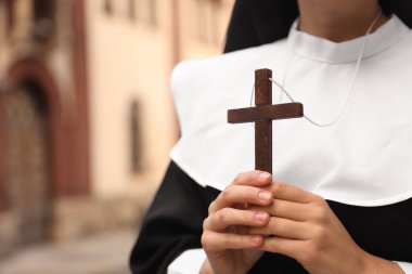 Young nun holding Christian cross near building outdoors, closeup. Space for text clipart