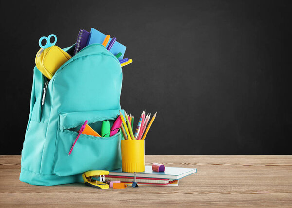 Backpack with school stationery on wooden table near black chalkboard, space for text