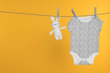Bebek tulumu ve tavşan oyuncağı çamaşır ipinde turuncu arka planda kuruyor, mesaj atmak için yer var.