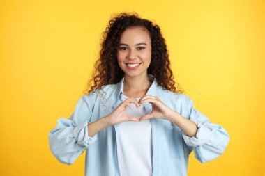 Happy young African-American woman making heart with hands on yellow background