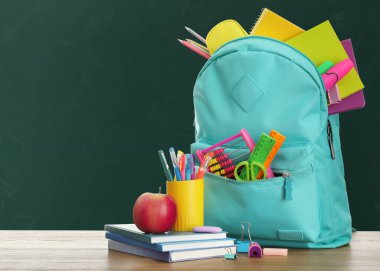 Yeşil karatahtanın yanındaki ahşap masanın üzerinde okul kırtasiyesi olan sırt çantası, metin için alan