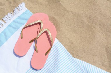 Beyaz ve açık mavi çizgili havlu, kumlu sahilde parmak arası terlikler, düz yatış. Metin için boşluk