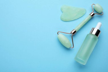 Jade gua sha aracı, yüz silindiri ve açık mavi arka planda bir şişe serum, düz konum. Metin için boşluk