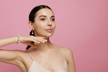 Pembe arka planda zarif inci takılar takan genç bir kadın.