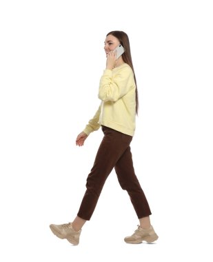 Sıradan kıyafetli genç bir kadın beyaz arka planda yürürken akıllı telefondan konuşuyor.