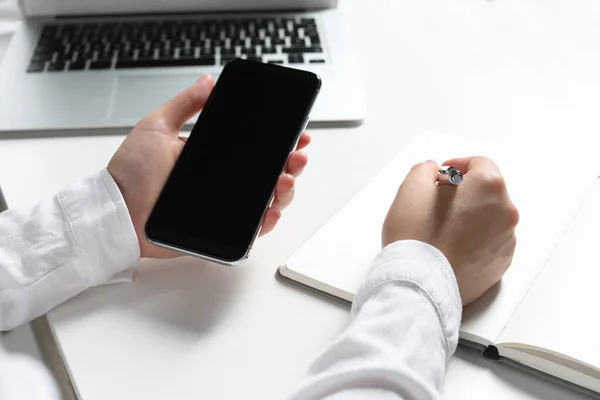 stock image Woman with smartphone writing in notebook at white table, closeup