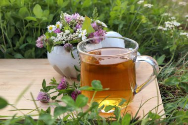 Bir fincan aromatik bitkisel çay, tokmak ve seramik havan, yeşil çimlerin üzerinde farklı kır çiçekleri.