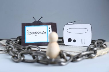 Propaganda concept. Human mired in media field. Chained wooden figure, newspapers, paper TV and radio on table clipart