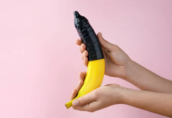 ピンクの背景にコンドームをつけてバナナを持っている女性 クローズアップ 安全なセックスの概念 — ストック写真