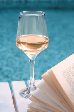 Glass of tasty wine and open book on wooden table near swimming pool clipart