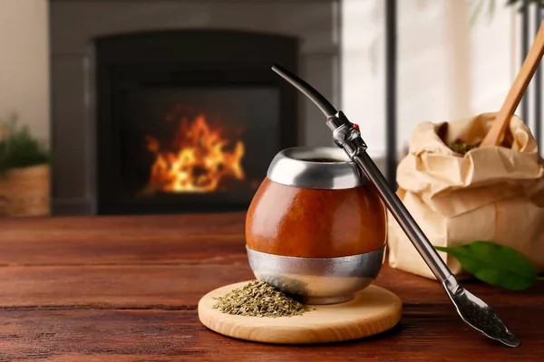 Calabash with mate tea and bombilla on wooden table near fireplace. Space for text