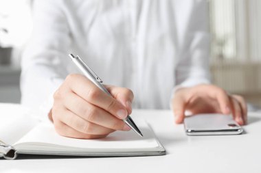 Ofisteki beyaz masada, defterine akıllı telefon yazan kadın, yakın plan.