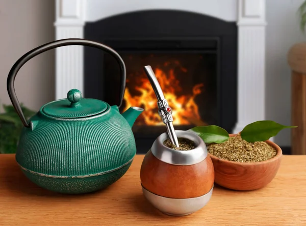 Calabash with mate tea, bombilla and teapot on wooden table near fireplace