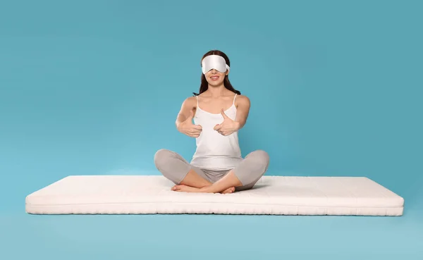 Woman in sleep mask sitting on soft mattress and showing thumbs up against light blue background