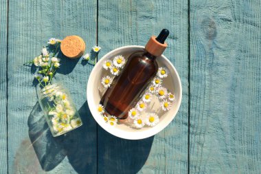 Bottle of chamomile essential oil and flowers on light blue wooden table, flat lay