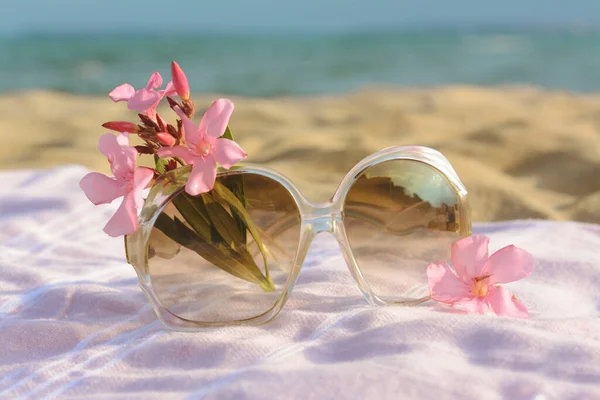 Beautiful sunglasses with tropical flower on blanket near sea, closeup