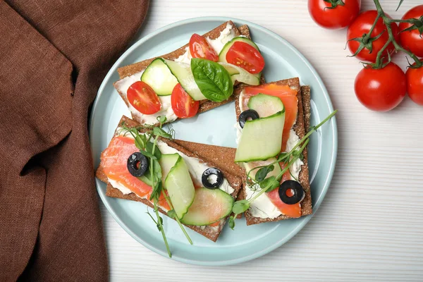 stock image Tasty rye crispbreads with salmon, cream cheese and vegetables served on white wooden table, flat lay