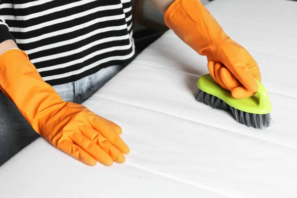 Woman in orange gloves cleaning white mattress with brush indoors, closeup