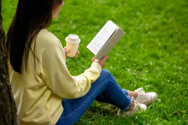 在公园里拿着一杯咖啡看书的年轻女人 — 图库照片