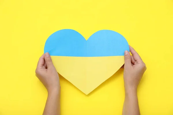 Woman holding paper heart in colors of Ukrainian flag on yellow background, top view