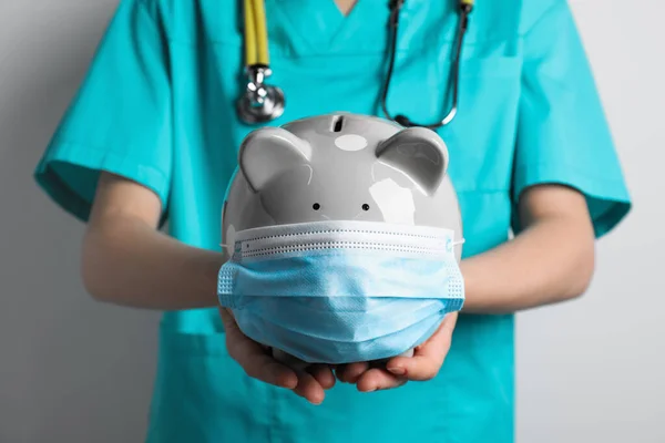 stock image Doctor holding light gray ceramic piggy bank with mask against white wall, closeup. Medical insurance