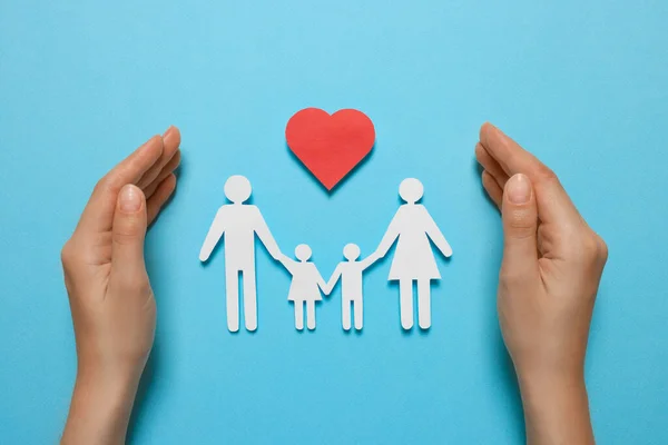 Woman protecting paper family figures and red heart on light blue background, top view. Insurance concept