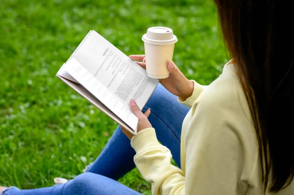 Young woman with cup of coffee reading book in park, closeup