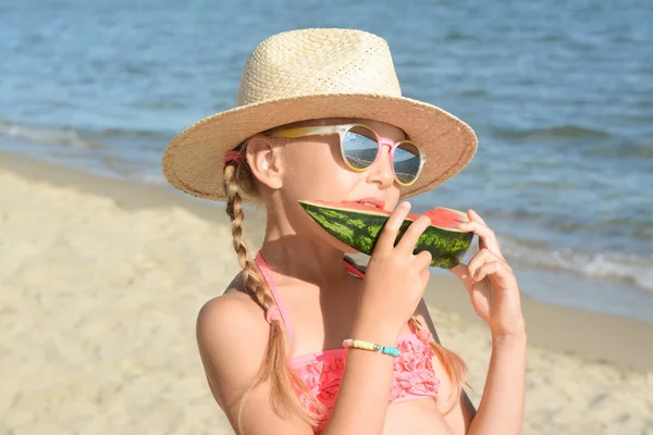 stock image Cute little girl in straw hat and sunglasses eating juicy watermelon on beach