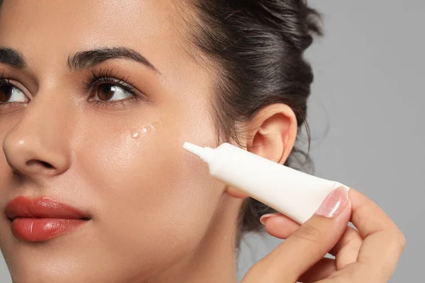 Woman applying cream under eyes on grey background, closeup. Skin care