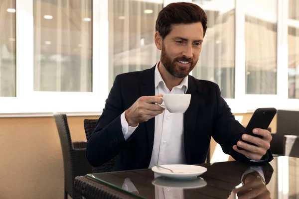 一个英俊的留着胡子的男人 在咖啡店的桌子旁 拿着一杯饮料和智能手机 — 图库照片