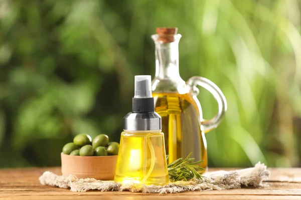 Bottles with cooking oil, olives and rosemary on wooden table against blurred background
