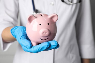 Doktor elinde açık pembe seramik domuz kumbarası tutuyor, yakın plan. Sağlık sigortası