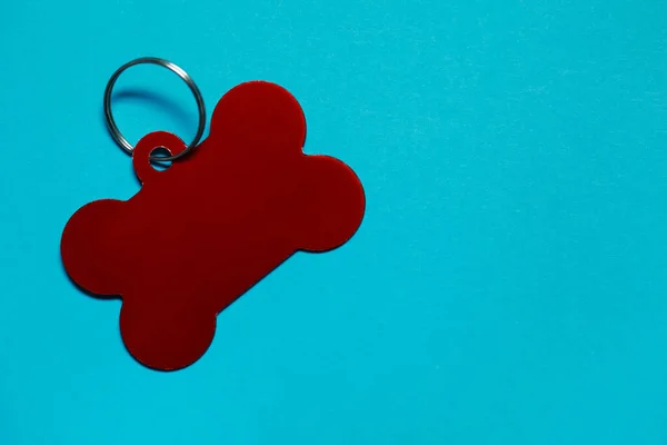 Red pet tag in shape of bone with ring on light blue background, top view. Space for text