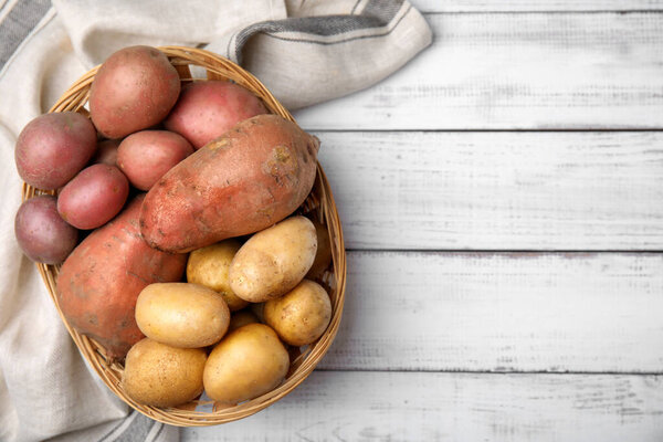 Different types of fresh potatoes in wicker basket on white wooden table, flat lay. Space for text
