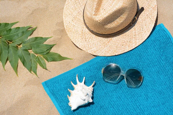 stock image Soft blue beach towel with sunglasses, straw hat, seashell and green leaves on sand, flat lay