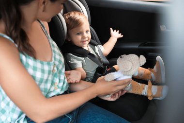 Woman with knitted toy near her daughter in child safety seat inside car clipart