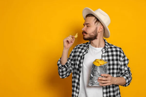 Handsome young man with bag of tasty potato chips on yellow background. Space for text