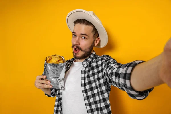 Handsome young man with bag of tasty potato chips taking selfie on yellow background