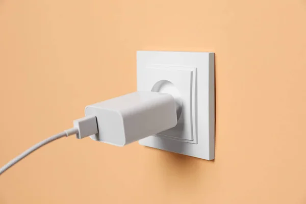 Charger adapter plugged into power socket on pale orange wall, closeup. Electrical supply
