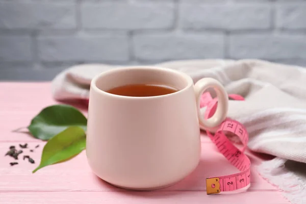 Cup of herbal diet tea and measuring tape on pink wooden table against brick wall, closeup. Weight loss concept