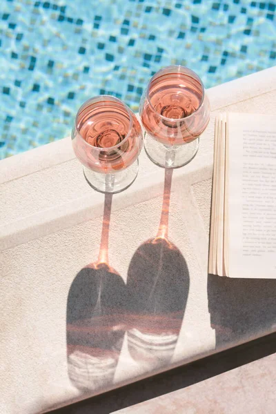 stock image Glasses of tasty rose wine and open book on swimming pool edge, above view